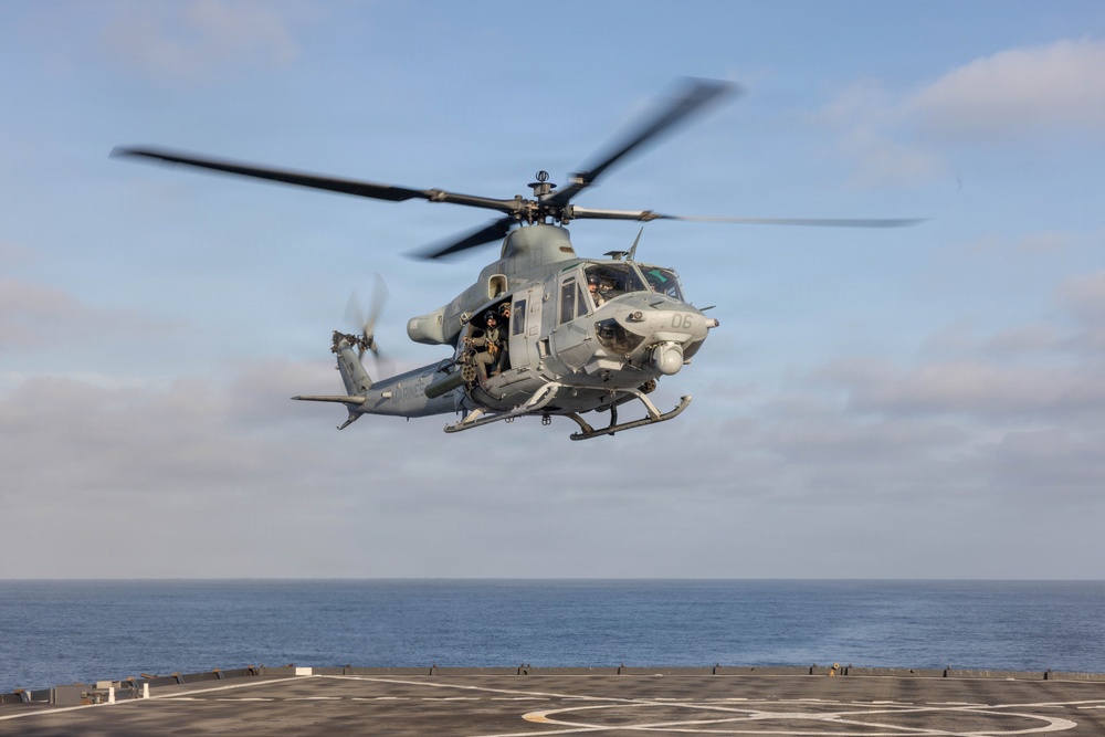 A military helicopter, identified by the number '06' on its nose, is hovering or taking off from a naval vessel's flight deck over open ocean. The aircraft appears to be a Bell UH-1Y Venom or similar variant in gray military livery, with at least two crew members visible. The image was taken in clear daylight conditions with calm seas in the background.