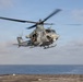 A military helicopter, identified by the number '06' on its nose, is hovering or taking off from a naval vessel's flight deck over open ocean. The aircraft appears to be a Bell UH-1Y Venom or similar variant in gray military livery, with at least two crew members visible. The image was taken in clear daylight conditions with calm seas in the background.