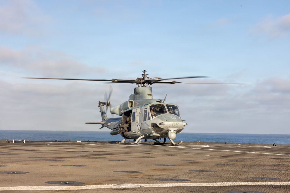 A military helicopter, identified as number '06', is positioned on the flight deck of a naval vessel at sea. The rotors are spinning, suggesting the aircraft is either preparing for takeoff or has just landed. The setting is an open ocean environment under partly cloudy skies.