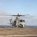 A military helicopter, identified as number '06', is positioned on the flight deck of a naval vessel at sea. The rotors are spinning, suggesting the aircraft is either preparing for takeoff or has just landed. The setting is an open ocean environment under partly cloudy skies.