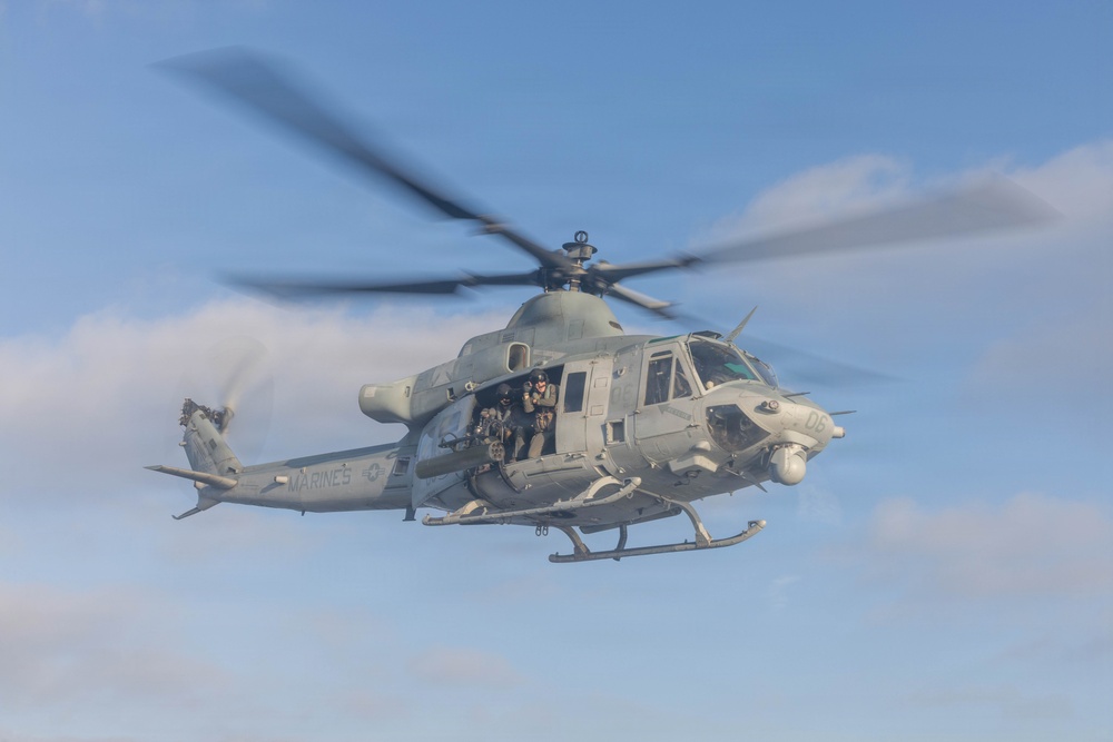 A U.S. Marines military helicopter is captured in flight against a partly cloudy blue sky. The aircraft, marked 'MARINES' on its fuselage with the identifier 'D6', has its side door open with armed personnel visible inside. The rotor blades show motion blur, indicating active flight.