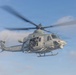 A U.S. Marines military helicopter is captured in flight against a partly cloudy blue sky. The aircraft, marked 'MARINES' on its fuselage with the identifier 'D6', has its side door open with armed personnel visible inside. The rotor blades show motion blur, indicating active flight.