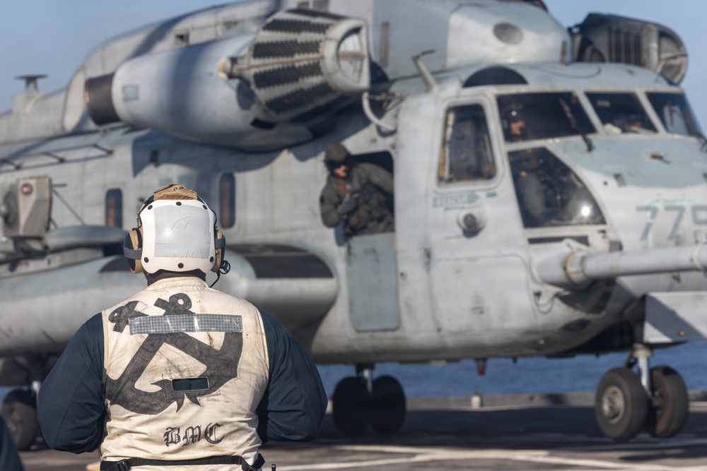 A flight deck crew member wearing a white vest with an anchor insignia and 'BMC' text stands on a ship's flight deck observing a large military helicopter. The helicopter, marked with the number '779', appears to be a CH-53 Sea Stallion or similar heavy-lift helicopter in a gray/blue military paint scheme. The scene takes place on what appears to be a naval vessel's flight deck, with crew members visible in and around the aircraft.