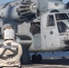 A flight deck crew member wearing a white vest with an anchor insignia and 'BMC' text stands on a ship's flight deck observing a large military helicopter. The helicopter, marked with the number '779', appears to be a CH-53 Sea Stallion or similar heavy-lift helicopter in a gray/blue military paint scheme. The scene takes place on what appears to be a naval vessel's flight deck, with crew members visible in and around the aircraft.