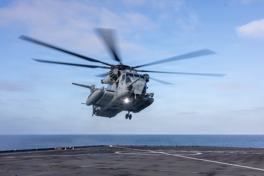 A large military helicopter is hovering or taking off from the flight deck of a naval vessel at sea. The helicopter appears to be a CH-53E Super Stallion, identifiable by its large tandem rotor configuration and gray military paint scheme. The image was captured with a relatively fast shutter speed showing rotor blur, over open ocean under partly cloudy skies.