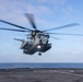 A large military helicopter is hovering or taking off from the flight deck of a naval vessel at sea. The helicopter appears to be a CH-53E Super Stallion, identifiable by its large tandem rotor configuration and gray military paint scheme. The image was captured with a relatively fast shutter speed showing rotor blur, over open ocean under partly cloudy skies.