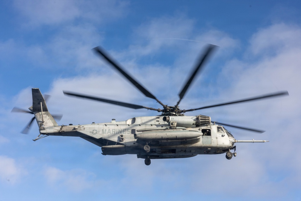 A large U.S. Marines heavy-lift helicopter is captured in flight against a partly cloudy blue sky. The aircraft is photographed head-on at close range, with its main rotor blades visibly blurred from rotation. The markings and configuration identify it as a Sikorsky CH-53E Super Stallion operated by the United States Marine Corps.