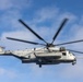 A large U.S. Marines heavy-lift helicopter is captured in flight against a partly cloudy blue sky. The aircraft is photographed head-on at close range, with its main rotor blades visibly blurred from rotation. The markings and configuration identify it as a Sikorsky CH-53E Super Stallion operated by the United States Marine Corps.
