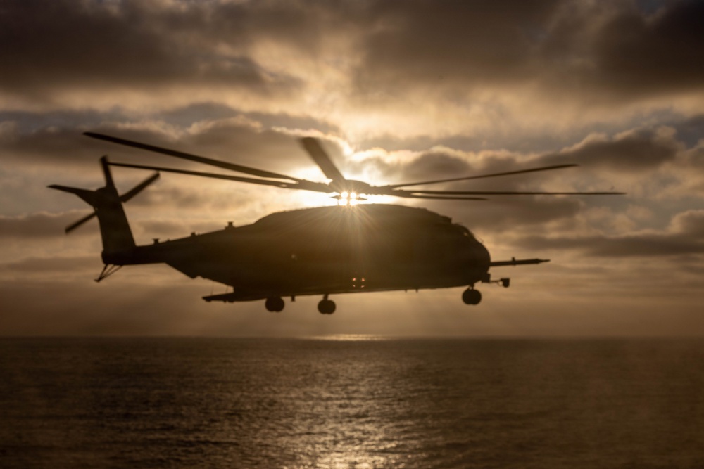 A large military helicopter is captured in silhouette against a dramatic sunset sky over open water. The sun bursts through clouds directly behind the aircraft, creating a strong backlit effect that obscures fine details. The scene conveys a sense of operational maritime activity in low-light conditions.