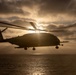 A large military helicopter is captured in silhouette against a dramatic sunset sky over open water. The sun bursts through clouds directly behind the aircraft, creating a strong backlit effect that obscures fine details. The scene conveys a sense of operational maritime activity in low-light conditions.