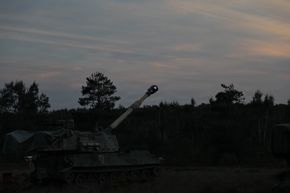 A self-propelled artillery vehicle is photographed at dusk in what appears to be a field or training area surrounded by trees. The vehicle's long gun barrel is elevated and pointed upward at an angle, with a distinctive muzzle brake visible at the tip. The low-light conditions create a silhouette effect against a twilight sky with wispy clouds.