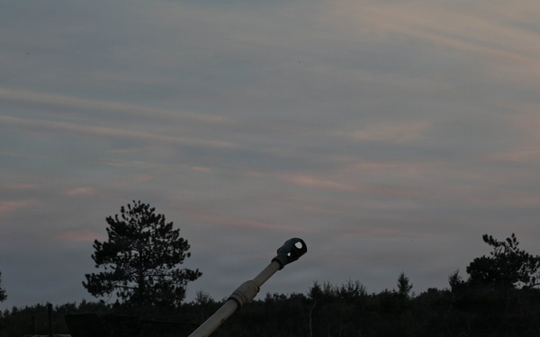 A self-propelled artillery vehicle is photographed at dusk in what appears to be a field or training area surrounded by trees. The vehicle's long gun barrel is elevated and pointed upward at an angle, with a distinctive muzzle brake visible at the tip. The low-light conditions create a silhouette effect against a twilight sky with wispy clouds.