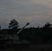 A self-propelled artillery vehicle is photographed at dusk in what appears to be a field or training area surrounded by trees. The vehicle's long gun barrel is elevated and pointed upward at an angle, with a distinctive muzzle brake visible at the tip. The low-light conditions create a silhouette effect against a twilight sky with wispy clouds.