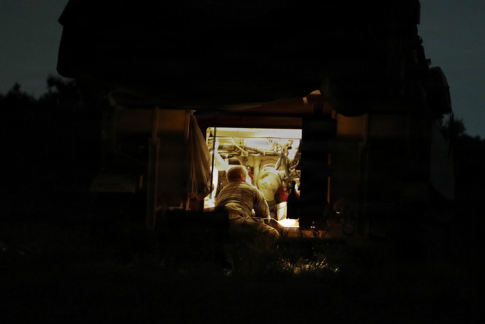 A nighttime photograph showing a person working inside or on a large vehicle or piece of machinery, illuminated by an artificial light source. The scene is viewed through a gap or opening between two large dark structures, creating a framed tunnel-like perspective. The surrounding environment is nearly completely dark, with only the work area lit.