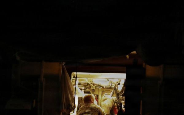 A nighttime photograph showing a person working inside or on a large vehicle or piece of machinery, illuminated by an artificial light source. The scene is viewed through a gap or opening between two large dark structures, creating a framed tunnel-like perspective. The surrounding environment is nearly completely dark, with only the work area lit.
