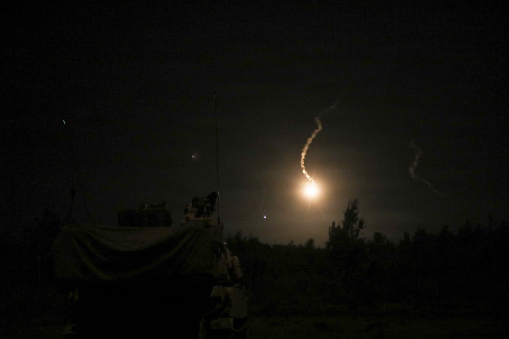 A nighttime image capturing what appears to be a missile or rocket launch, with a bright glowing exhaust trail visible against the dark sky. A military vehicle or equipment is silhouetted in the foreground on the left side. The scene is set near a treeline, suggesting a field or open terrain environment.