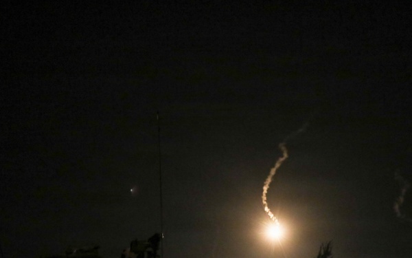 A nighttime image capturing what appears to be a missile or rocket launch, with a bright glowing exhaust trail visible against the dark sky. A military vehicle or equipment is silhouetted in the foreground on the left side. The scene is set near a treeline, suggesting a field or open terrain environment.