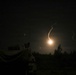 A nighttime image capturing what appears to be a missile or rocket launch, with a bright glowing exhaust trail visible against the dark sky. A military vehicle or equipment is silhouetted in the foreground on the left side. The scene is set near a treeline, suggesting a field or open terrain environment.