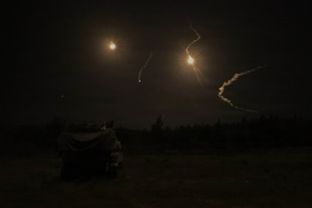 A nighttime outdoor scene showing multiple bright light sources in the sky with visible smoke or vapor trails, consistent with rocket or missile launches or aerial munitions. The ground level shows a dark treeline silhouette and what appears to be a partially visible vehicle or equipment on the left foreground. The long-exposure photograph captures the trajectories of at least two projectiles with glowing exhaust plumes.