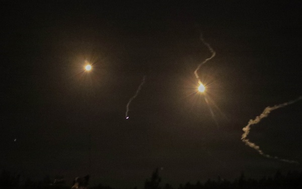 A nighttime outdoor scene showing multiple bright light sources in the sky with visible smoke or vapor trails, consistent with rocket or missile launches or aerial munitions. The ground level shows a dark treeline silhouette and what appears to be a partially visible vehicle or equipment on the left foreground. The long-exposure photograph captures the trajectories of at least two projectiles with glowing exhaust plumes.