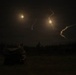 A nighttime outdoor scene showing multiple bright light sources in the sky with visible smoke or vapor trails, consistent with rocket or missile launches or aerial munitions. The ground level shows a dark treeline silhouette and what appears to be a partially visible vehicle or equipment on the left foreground. The long-exposure photograph captures the trajectories of at least two projectiles with glowing exhaust plumes.