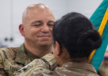 Two U.S. Army soldiers are engaged in what appears to be a military change of command or award ceremony. The soldier facing the camera, identified by the name tape 'LIVER' (partially visible), is smiling while the other soldier appears to be pinning something or presenting an item. A teal/turquoise guidon with gold fringe is visible in the background.
