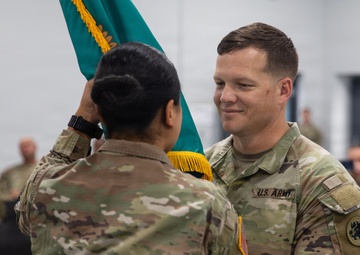 A U.S. Army change of command ceremony is taking place in an indoor military facility. Two soldiers in OCP (Operational Camouflage Pattern) uniforms are exchanging a teal/green guidon flag, which is the central symbolic act of a change of command. A formation of seated soldiers in uniform is visible in the background observing the ceremony.