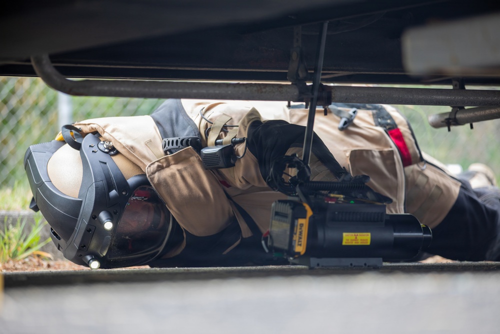 A person in tan/khaki tactical or bomb disposal gear is lying prone on the ground, inspecting the underside of a vehicle. They are equipped with a specialized helmet, radio/communication device, and are holding what appears to be a DeWalt-branded inspection or detection tool. The scene suggests a security, law enforcement, or explosive ordnance disposal (EOD) operation.