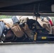 A person in tan/khaki tactical or bomb disposal gear is lying prone on the ground, inspecting the underside of a vehicle. They are equipped with a specialized helmet, radio/communication device, and are holding what appears to be a DeWalt-branded inspection or detection tool. The scene suggests a security, law enforcement, or explosive ordnance disposal (EOD) operation.