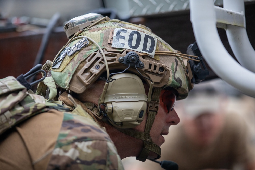 A close-up profile shot of a military EOD (Explosive Ordnance Disposal) technician wearing a heavily equipped tactical helmet with multiple attachments. The individual is wearing OCP/MultiCam camouflage and red-tinted protective eyewear. The image captures significant detail of the helmet-mounted equipment and unit identification markings.