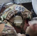 A close-up profile shot of a military EOD (Explosive Ordnance Disposal) technician wearing a heavily equipped tactical helmet with multiple attachments. The individual is wearing OCP/MultiCam camouflage and red-tinted protective eyewear. The image captures significant detail of the helmet-mounted equipment and unit identification markings.