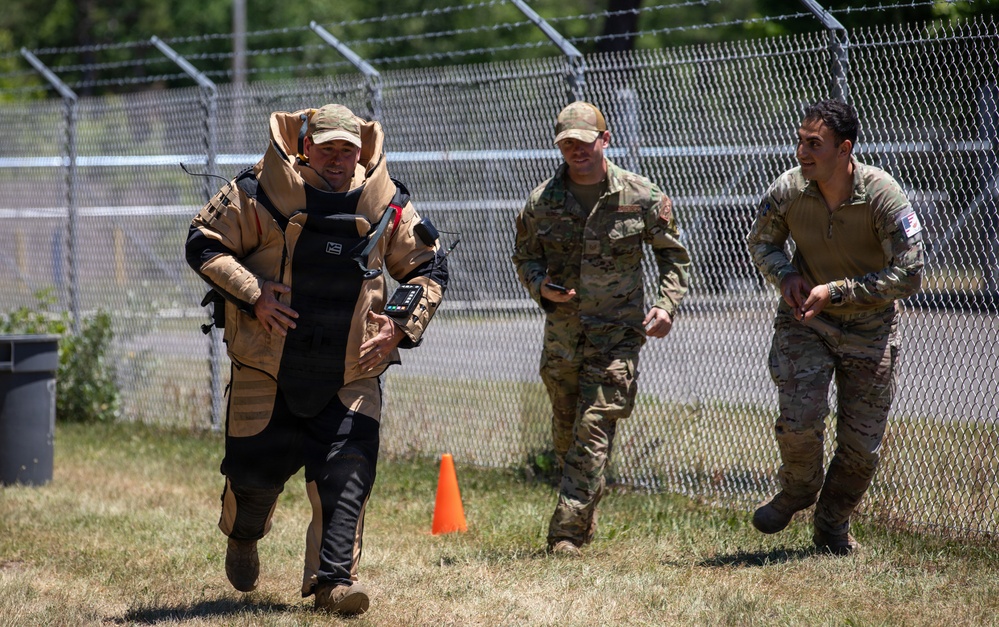 Three military personnel are walking along a chain-link fence topped with barbed wire in an outdoor training area. The person on the left is wearing an EOD (Explosive Ordnance Disposal) bomb suit in tan and black, while the other two are in standard multicam/OCP military uniforms. The atmosphere appears relaxed, with the two uniformed soldiers smiling.