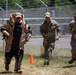 Three military personnel are walking along a chain-link fence topped with barbed wire in an outdoor training area. The person on the left is wearing an EOD (Explosive Ordnance Disposal) bomb suit in tan and black, while the other two are in standard multicam/OCP military uniforms. The atmosphere appears relaxed, with the two uniformed soldiers smiling.
