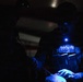 A low-light image showing what appears to be military or law enforcement personnel conducting a close-up technical task in near-darkness. Blue illumination from a mounted light source reveals tactical gear with 'BRAVE EOD' stenciled text, suggesting Explosive Ordnance Disposal personnel. The operators appear to be examining or manipulating an object on a surface using focused lighting.