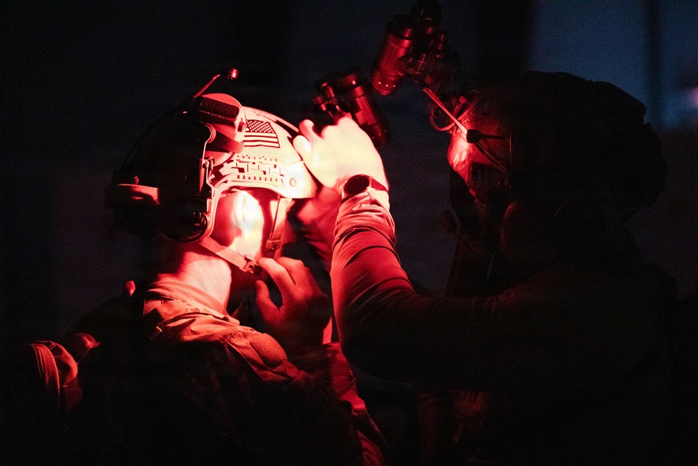 A nighttime military scene illuminated by red light showing two personnel in tactical gear. One individual wearing a helmet with a US flag patch and night vision device mount appears to be receiving medical attention or equipment adjustment from a second person. The red lighting suggests a low-visibility operational environment consistent with tactical night operations.
