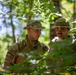 Two military personnel in camouflage uniforms and boonie hats are positioned among dense forest vegetation. They appear to be engaged in a field training exercise or reconnaissance activity, with one individual appearing to speak or brief the other. The image is captured through foliage, suggesting a naturalistic or candid perspective.