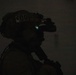 A silhouetted figure wearing tactical military/law enforcement gear is photographed in very low-light conditions. The individual is equipped with a helmet marked 'EOD' (Explosive Ordnance Disposal) and night vision goggles that emit a faint blue-white light. The image is predominantly dark, with the subject visible only in profile silhouette.