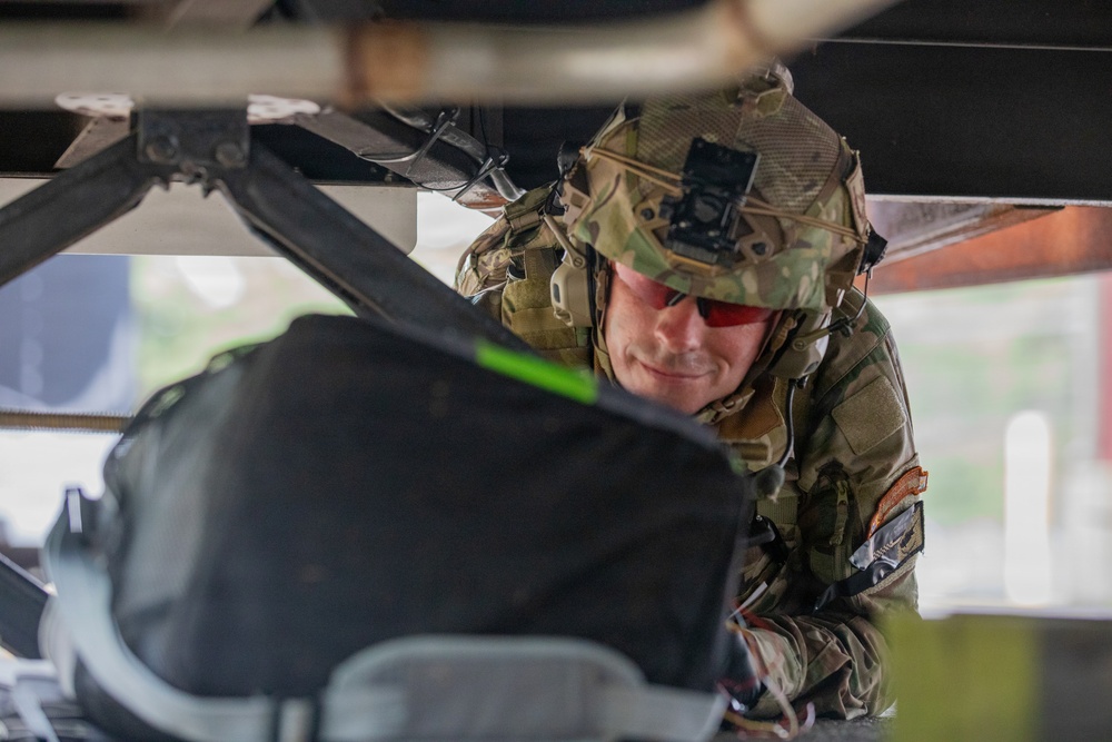 A soldier in full combat gear, wearing a camouflage uniform and helmet with a mounted device, is leaning forward to inspect or work on equipment beneath a large vehicle or aircraft structure. The soldier is wearing red-tinted protective eyewear and has unit patches visible on the uniform sleeve. The image is taken from a low angle, emphasizing the confined working space.