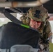 A soldier in full combat gear, wearing a camouflage uniform and helmet with a mounted device, is leaning forward to inspect or work on equipment beneath a large vehicle or aircraft structure. The soldier is wearing red-tinted protective eyewear and has unit patches visible on the uniform sleeve. The image is taken from a low angle, emphasizing the confined working space.