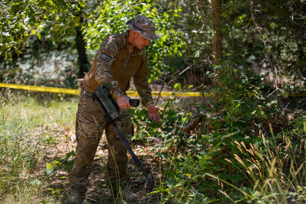 A military or law enforcement personnel in multicam/OCP camouflage uniform is using a handheld detection device, likely a metal detector or explosive ordnance detection instrument, in a wooded area. The individual is leaning forward and scanning vegetation and ground cover near what appears to be a marked perimeter indicated by yellow tape. The scene suggests a search, sweep, or EOD (Explosive Ordnance Disposal) training or operational activity.