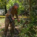 A military or law enforcement personnel in multicam/OCP camouflage uniform is using a handheld detection device, likely a metal detector or explosive ordnance detection instrument, in a wooded area. The individual is leaning forward and scanning vegetation and ground cover near what appears to be a marked perimeter indicated by yellow tape. The scene suggests a search, sweep, or EOD (Explosive Ordnance Disposal) training or operational activity.