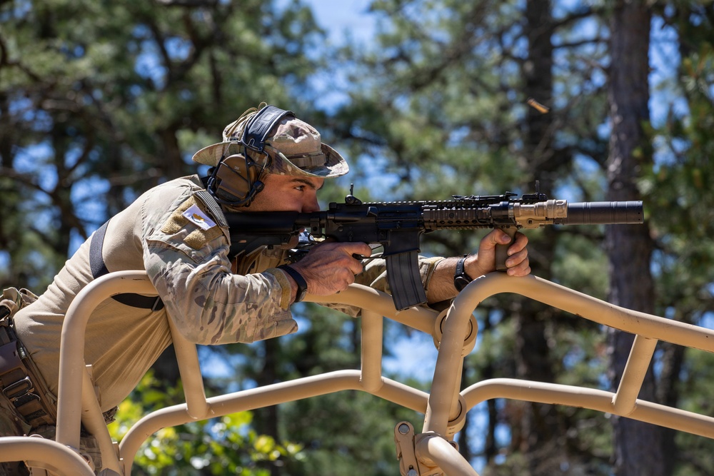 A soldier in camouflage uniform is firing a rifle from an elevated position on what appears to be a military vehicle roll cage. The shooter is bracing against the tan-colored tubular frame structure while aiming downrange. A spent brass casing is visible being ejected from the weapon, indicating active firing.