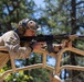 A soldier in camouflage uniform is firing a rifle from an elevated position on what appears to be a military vehicle roll cage. The shooter is bracing against the tan-colored tubular frame structure while aiming downrange. A spent brass casing is visible being ejected from the weapon, indicating active firing.