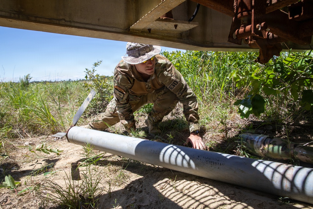 A soldier in multicam/OCP camouflage uniform and boonie hat is crouched beneath a structure, examining or handling what appears to be a large cylindrical tube or pipe on the ground. The setting is an outdoor rural or field environment with sandy soil, green vegetation, and what appears to be a rusted metal structure overhead. The scene suggests an EOD (Explosive Ordnance Disposal), inspection, or training activity.