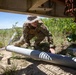 A soldier in multicam/OCP camouflage uniform and boonie hat is crouched beneath a structure, examining or handling what appears to be a large cylindrical tube or pipe on the ground. The setting is an outdoor rural or field environment with sandy soil, green vegetation, and what appears to be a rusted metal structure overhead. The scene suggests an EOD (Explosive Ordnance Disposal), inspection, or training activity.