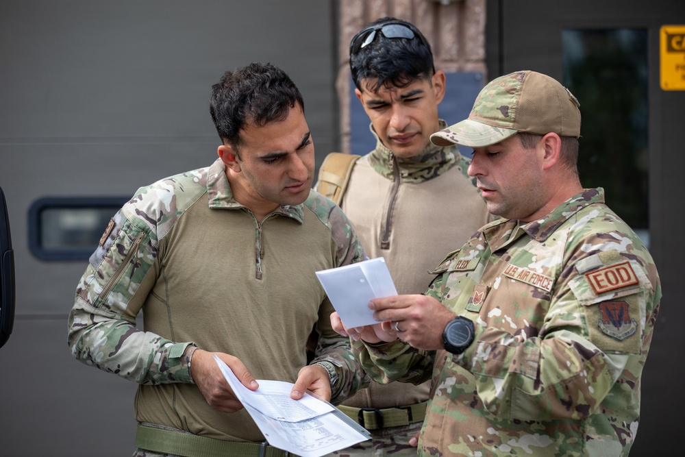 Three military personnel in MultiCam/OCP camouflage uniforms are reviewing documents together outdoors. The individual on the right is identifiable as a U.S. Air Force EOD (Explosive Ordnance Disposal) technician based on visible patches and insignia. The group appears to be conducting a briefing or reviewing operational paperwork.