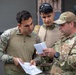 Three military personnel in MultiCam/OCP camouflage uniforms are reviewing documents together outdoors. The individual on the right is identifiable as a U.S. Air Force EOD (Explosive Ordnance Disposal) technician based on visible patches and insignia. The group appears to be conducting a briefing or reviewing operational paperwork.