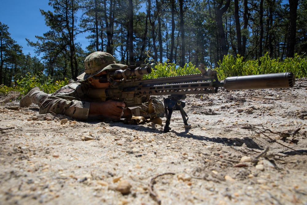 A military service member in full multicam camouflage uniform is in a prone shooting position on sandy ground, aiming a large precision rifle equipped with a suppressor, bipod, and optical scope. The setting appears to be an outdoor training range surrounded by pine trees under a clear blue sky. The image is taken from a low ground-level angle, emphasizing the weapon system in the foreground.