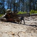 A military service member in full multicam camouflage uniform is in a prone shooting position on sandy ground, aiming a large precision rifle equipped with a suppressor, bipod, and optical scope. The setting appears to be an outdoor training range surrounded by pine trees under a clear blue sky. The image is taken from a low ground-level angle, emphasizing the weapon system in the foreground.