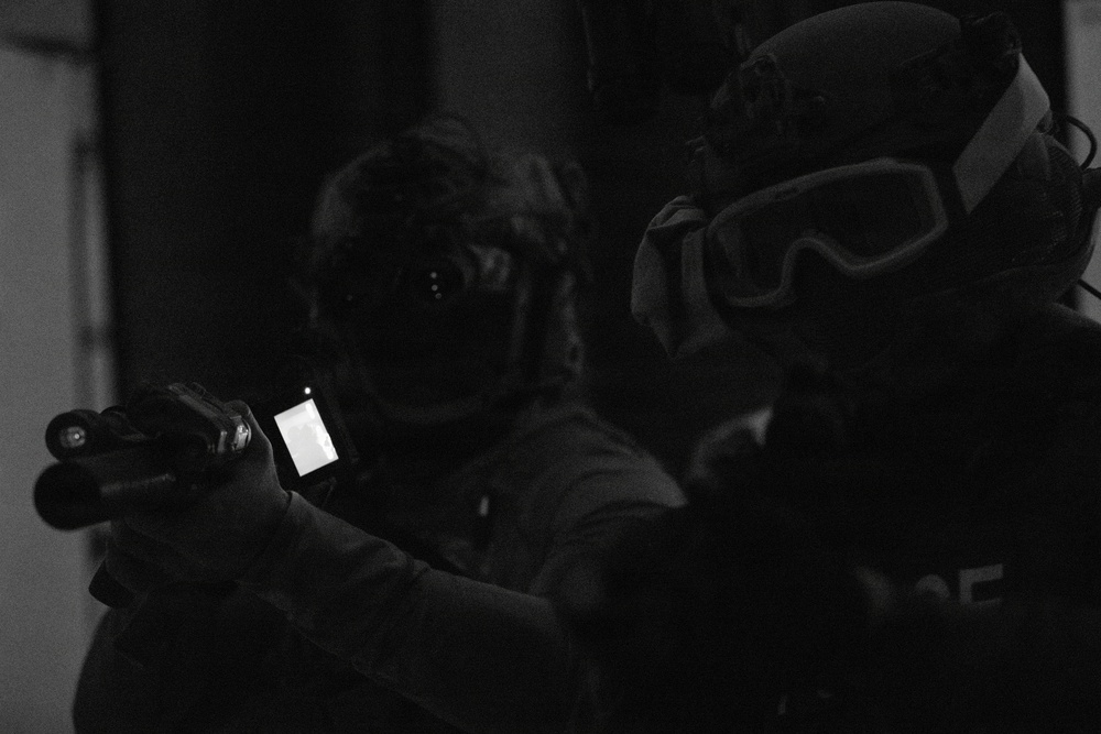 A black and white low-light photograph showing two individuals in tactical gear operating in a dark environment. One person is holding what appears to be a firearm with an attached illuminated device, possibly a weapon-mounted light or targeting system. Partial text visible on one individual's vest suggests law enforcement or tactical unit affiliation.